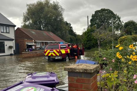 A fire and rescue vehicle parked in the middle of a flooded road, with water completely covering the width of the road and the gardens on one side. Wheelie bins and flowers are close by.