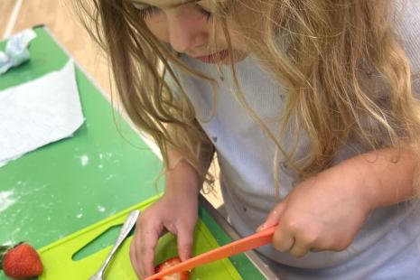 A young girl cutting a strawberry into slices with an orange knife on a green chopping board