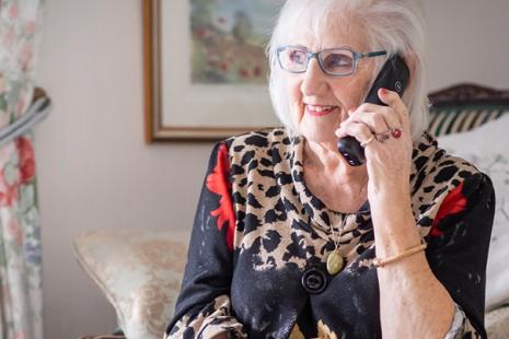 An older woman wearing glasses is sitting down and holding a phone to her ear.