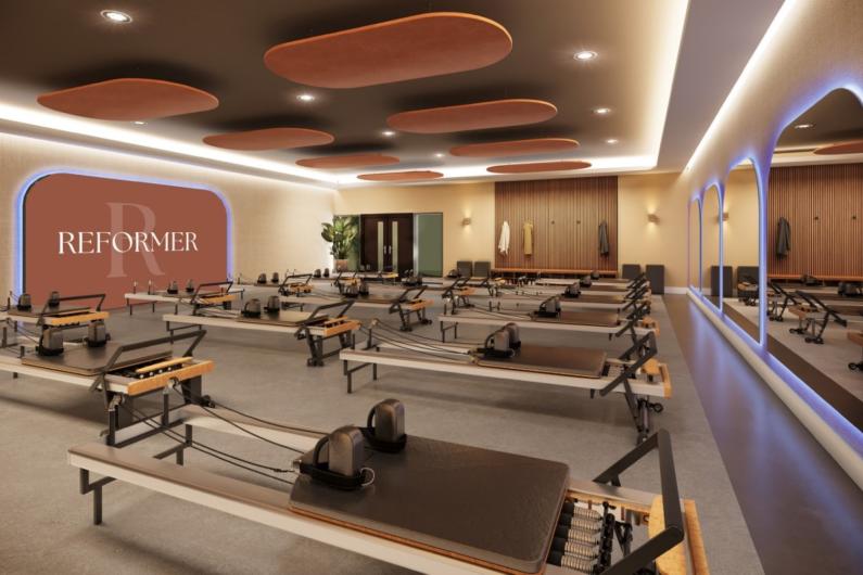 A CGI representation of the Reformer Pilates studio, with equipment arranged around the room with backlit mirrors at one end and a large pinkish backlit feature at the other end. Double doors and a large plant sit alongside a bench along the main wall, with coat hooks above.