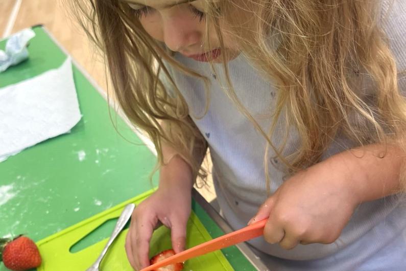 A young girl cutting a strawberry into slices with an orange knife on a green chopping board