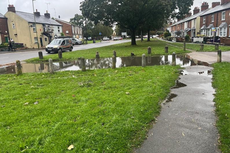 Woolgrove Road, with water covering a driveway and a few puddles on a grass verge, but the main road is clear.