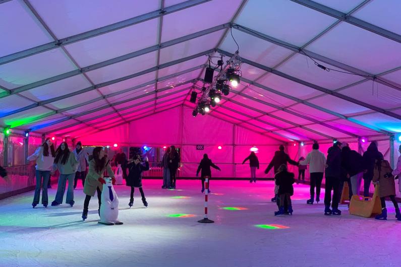 People skating round an ice rink that is lit up with bright colours, mostly pink and purple.