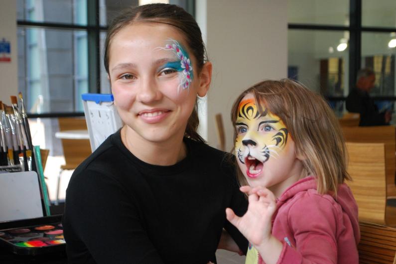 Hermione with a flower pattern painted on her face with a younger girl with a tiger painted on her face and pretending to growl with her mouth and hand