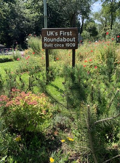 A sign on a roundabout saying 'UK's first roundabout built circa 1909' surrounded by different plants