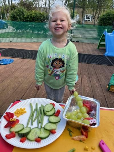 A little girl smiling widely with her plates of healthy food including cucumber and grapes